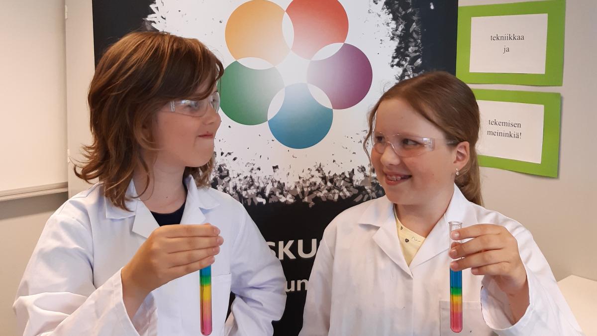 Kaksi lasta pitelevät koeputkia, joihin on tehty sokerisateenkaari ja hymyilevät toisilleen SAGA-tiedeluokassa.