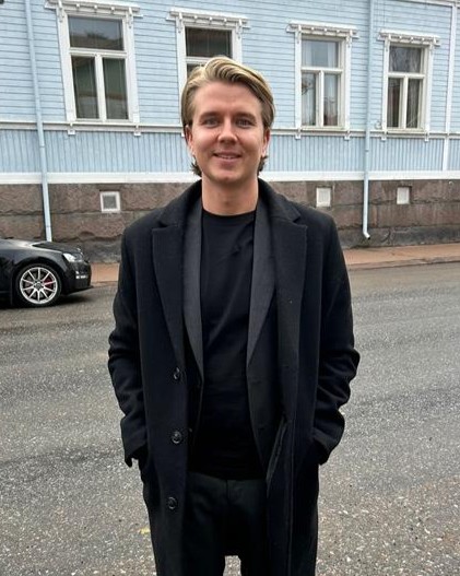 Mieshenkilö seisomassa tien vieressä. Hän poseeraa kameraan päin ja hymyilee. Kuvan taustalla näkyy sininen puutalo sekä mustan auton etuosa.