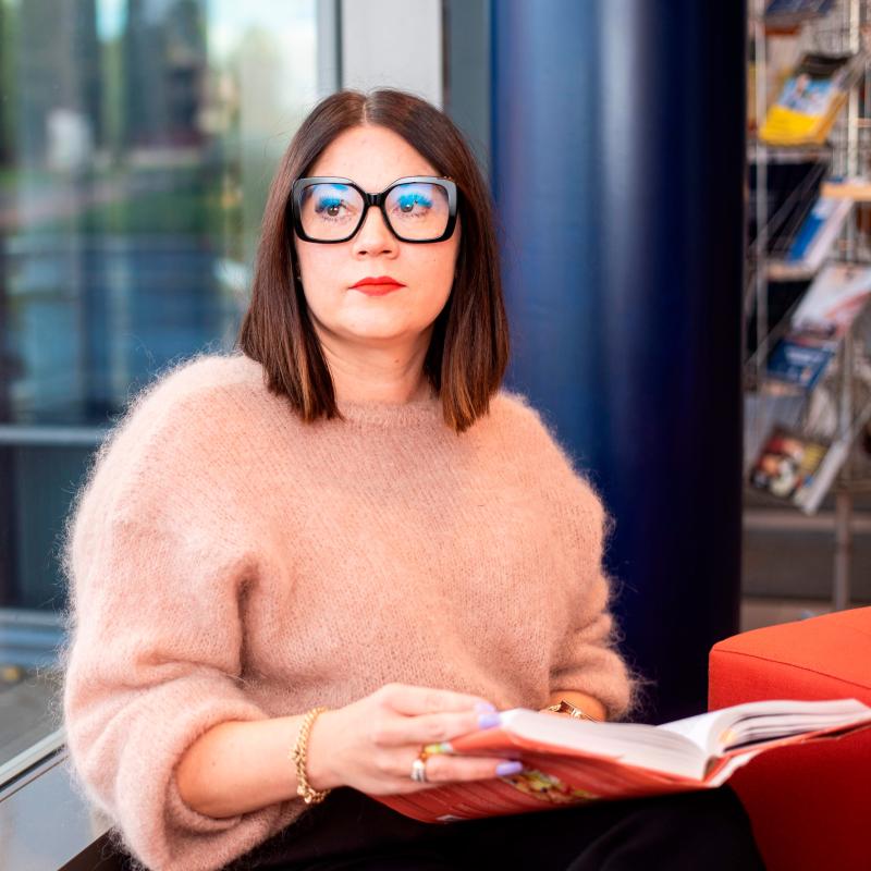 Naishenkilö istumassa pienellä sohvalla. Taustalla näkyy sumeana ikkuna. Naisella on päällä roosan värinen pörröinen neule ja silmillään hänellä on mustasankaiset silmälasit. Käsissään hän pitää kirjaa.