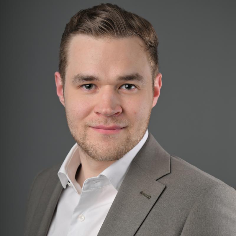 Mieshenkilö katsoo kameraan kasvoista otetussa lähikuvassa. Päällään hänellä on valkoinen kauluspaita ja ruskeanharmaa puvuntakki.