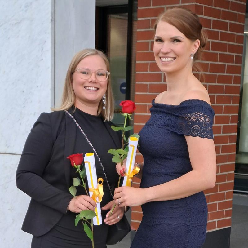 Kaksi henkilöä seisomassa juhlavaatteet päällä pihalla. Molemmat poseeraavat kameralle ja hymyilevät leveästi. Käsissään heillä molemmilla on paperikäärö sekä ruusu.