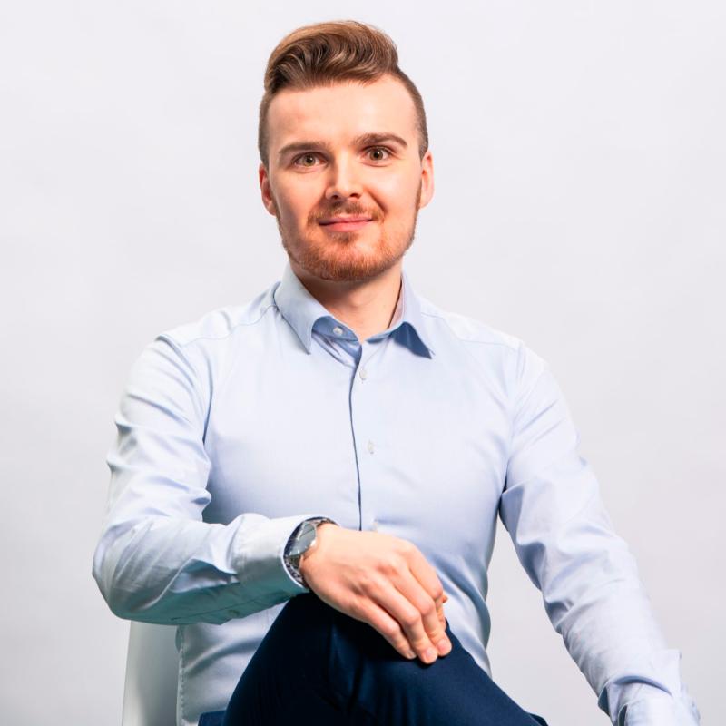 Mieshenkilö istumassa valkoisella tuolilla ja poseeraamassa kameraan. Hänellä on päällään vaalean sininen kauluspaita ja tummansiniset siistit housut. Hiukset ja silmät ovat ruskean väriset. Hän katsoo kohti kameraa ja hymyilee.