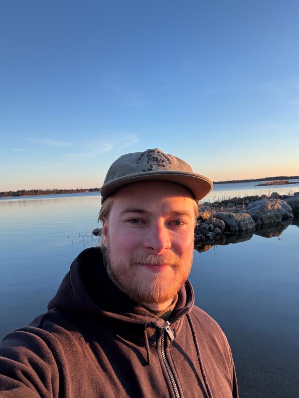 Mieshenkilö seisomassa meren rannassa. Hän poseeraa kameraa kohti ja taustalla näkyy tyyni vesi sekä kirkas sininen taivas-