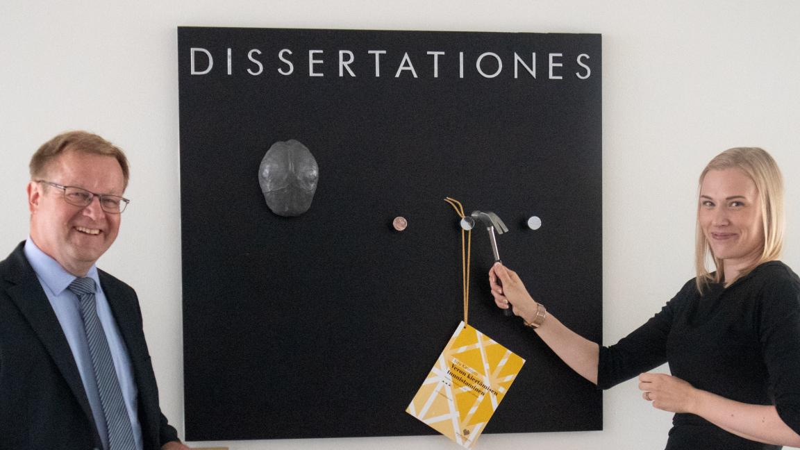 Siru Kaunisto and Juha Lindgren are about to nail a copy of Siru’s doctoral dissertation to the black Dissertationes board.