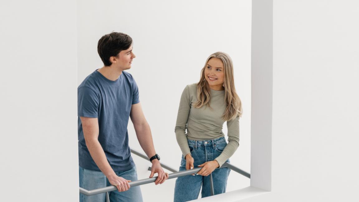 Poika ja tyttö nojaavat porrastasanteella kaiteeseen ja juttelevat keskenään