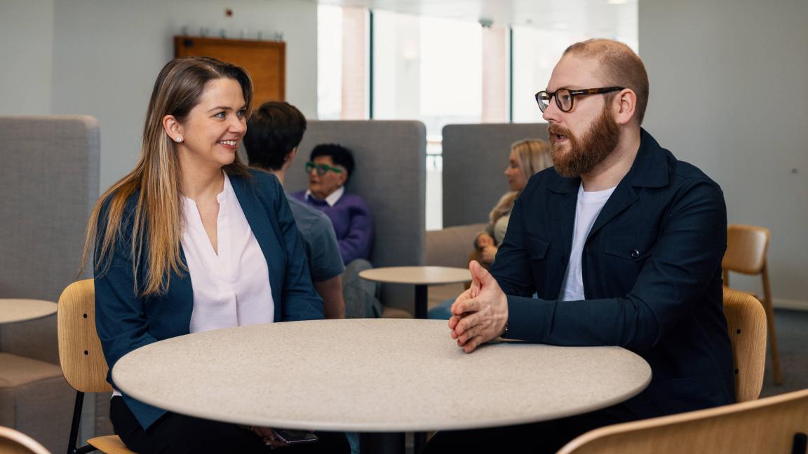 Kuvituskuva, kaksi henkilöä keskustelee pöydän ääressä.