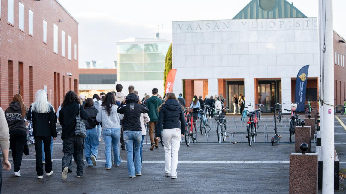 Useita henkiöitä kävelemässä kohti rakennusta. Rakennuksen yläreunassa lukee teksti "Vaasan yliopisto".