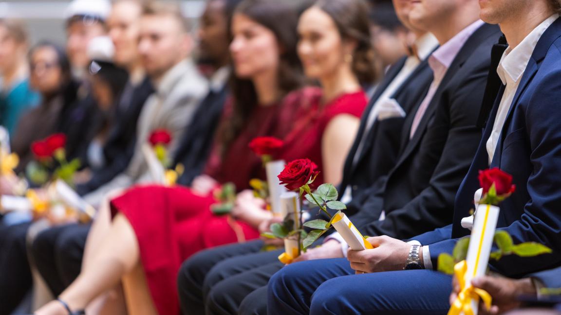 Valmistuneita opiskelijoita istumassa audiotriossa. Kuvan fokus on keskittnyt mieshenkilön kädessä olevaan ruusuun sekä muistaiskääröön. Taustalla näkyy sumeana muita henkilöitä.