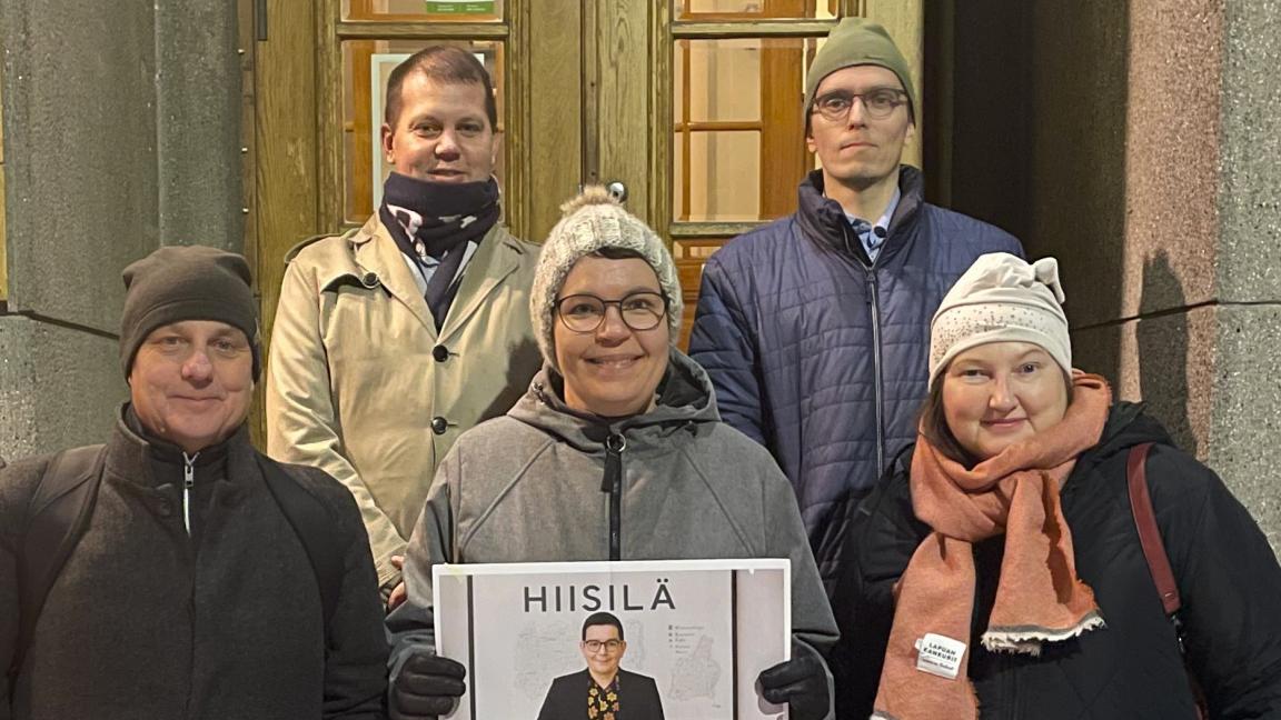 Viisi henkilöä seisomassa rakennuksen portailla kahdessa eri rivissä. Kaikki katsovat kameraan ja hymyilevät. Eturivissä keskellä olevalla henkilöllä on kädessään paperi, jossa on tekoälyllä luotu opettajan kuva kaikista portailla seisovista henkilöistä.