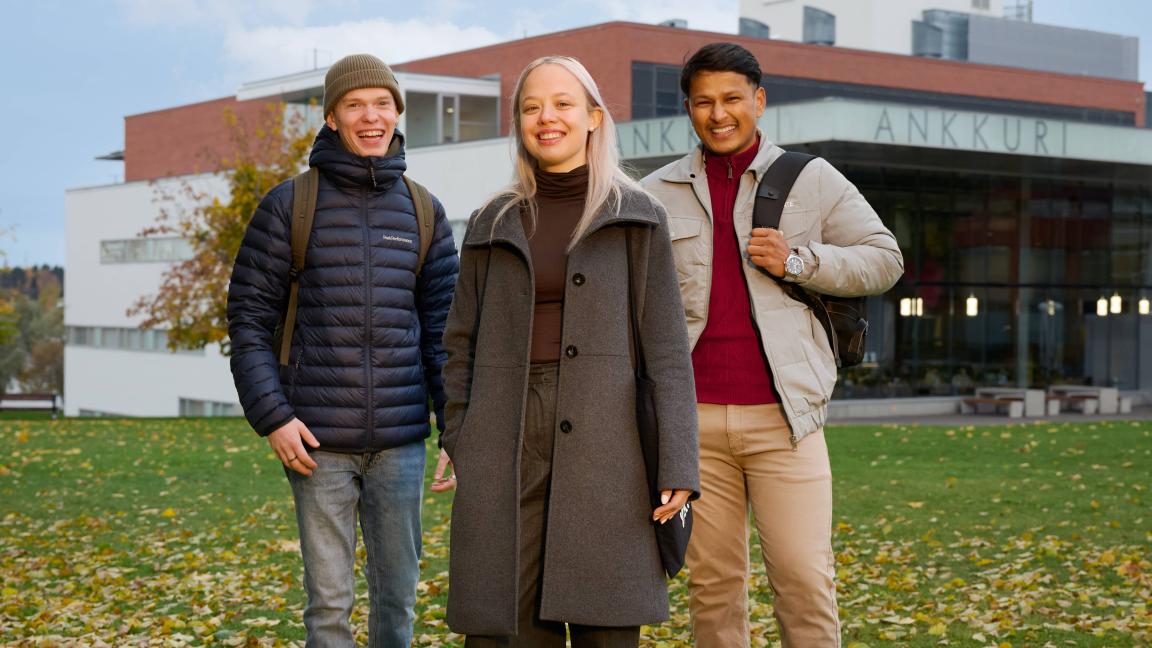 Kolme henkilöä seisomassa Vaasan yliopiston Ankkuri-rakennuksen edessä. Taustalla näkyy rakennus ja pilvinen sinitaivas.