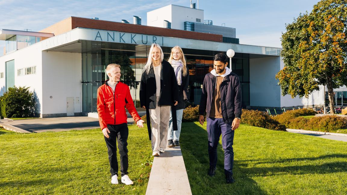 Neljä opiskelijaa kävelemässä aurinkoisella ja vehreällä yliopiston kampuksella. Kaksi opiskelijoista kävelee pienen korokkeen päällä, toiset kaksi kävelevät nurmella. Kaikilla on kasvoillaan ilmoinen ilme.