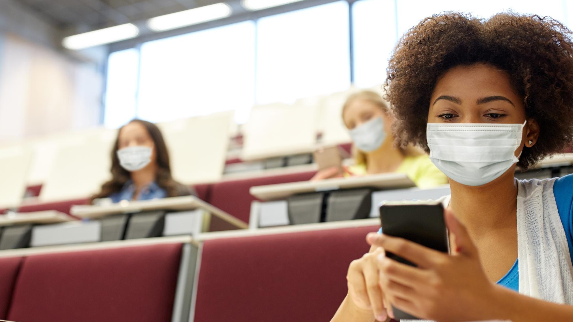 Nainen istuu luentosalissa kasvomaski kasvoilla puhelin kädessä. 