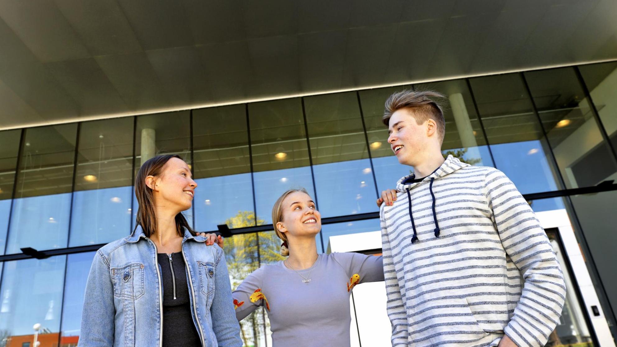 Kolme hymyilevää opiskelijaa seisoo yliopiston kampuksella