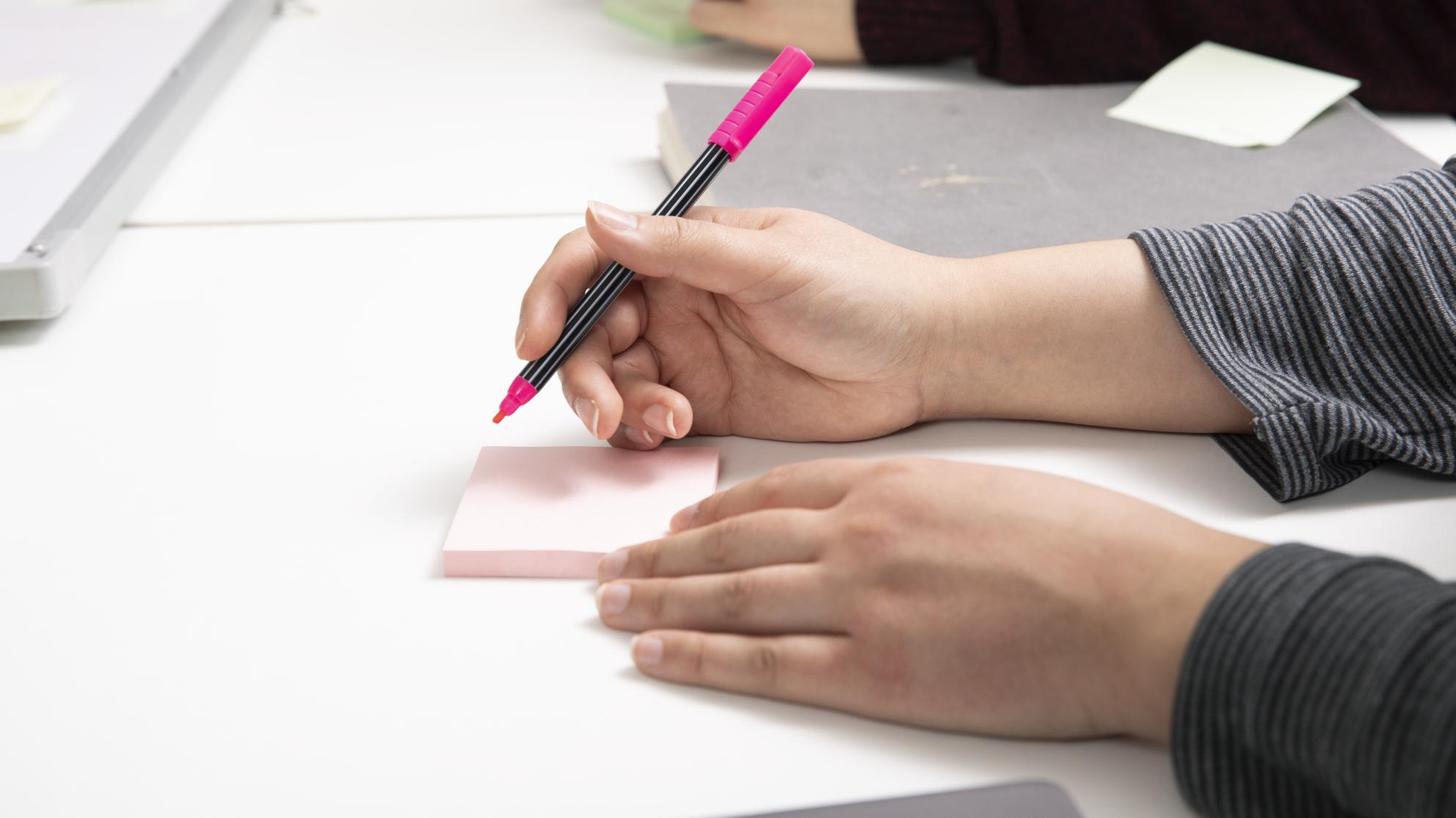 Kuvassa näkyy kädet, henkilö kirjoittaa post-it-lapulle