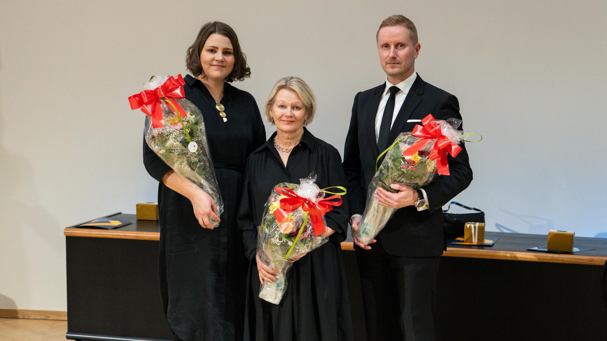 Kolme henkilöä seisomassa kukka-asetelmat käsissään. Kaikki katsovat kameraan ja heillä on päällään juhlavaatteet.