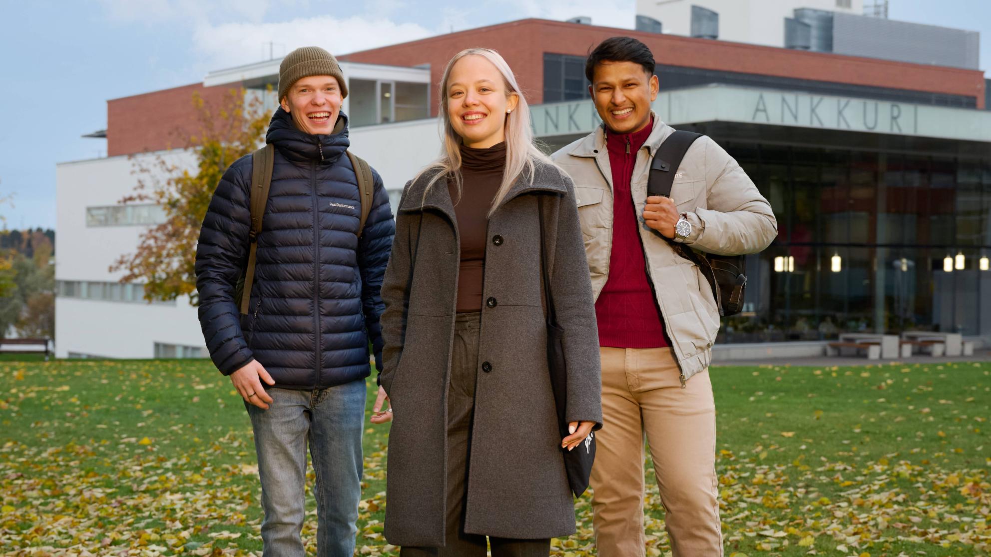 Kolme henkilöä seisomassa Vaasan yliopiston Ankkuri-rakennuksen edessä. Taustalla näkyy rakennus ja pilvinen sinitaivas.