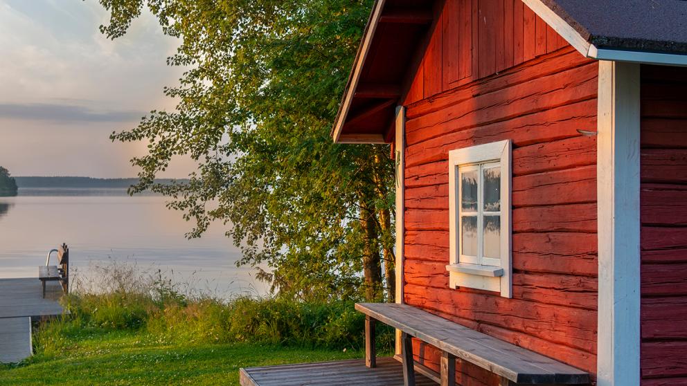 Mökkimaisema, jossa sauna on etualalla ja taustalla näkyy järvi.