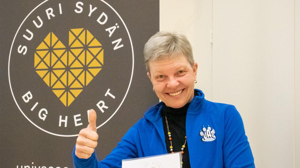 Tiina Jokinen peukuttaa hymyillen Vaasan yliopiston Suuri Sydän -bannerin edessä.