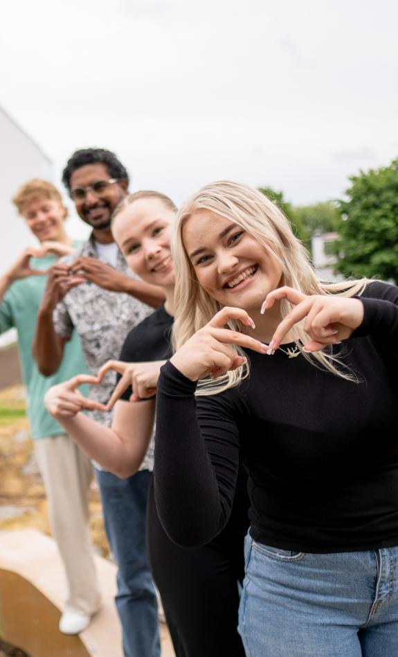 Neljä henkilöä poseeraa ja katsoo kameraan. Kaikki tekevät sormillaan sydämen ja hymyilevät isosti.