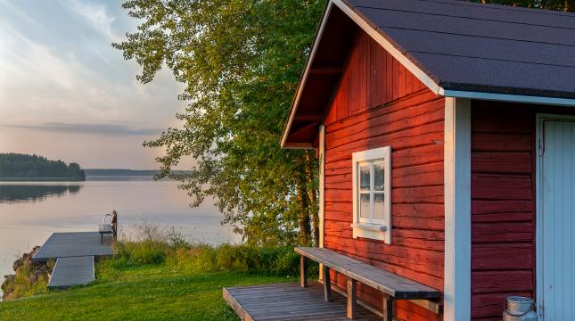 Mökkimaisema, jossa sauna on etualalla ja taustalla näkyy järvi.