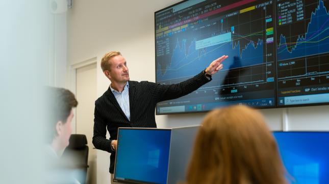 Opettaja osoittaa taululle, jossa pörssikursseja. 