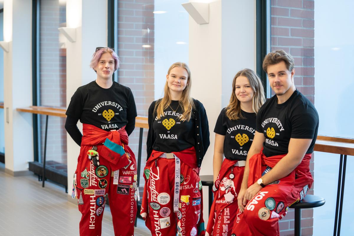Neljä opiskelijaa punaiset haalarit jalassa ja yliopiston musta t-paita päällä. Kaikki katsovat kameraan ja hymyilevät.