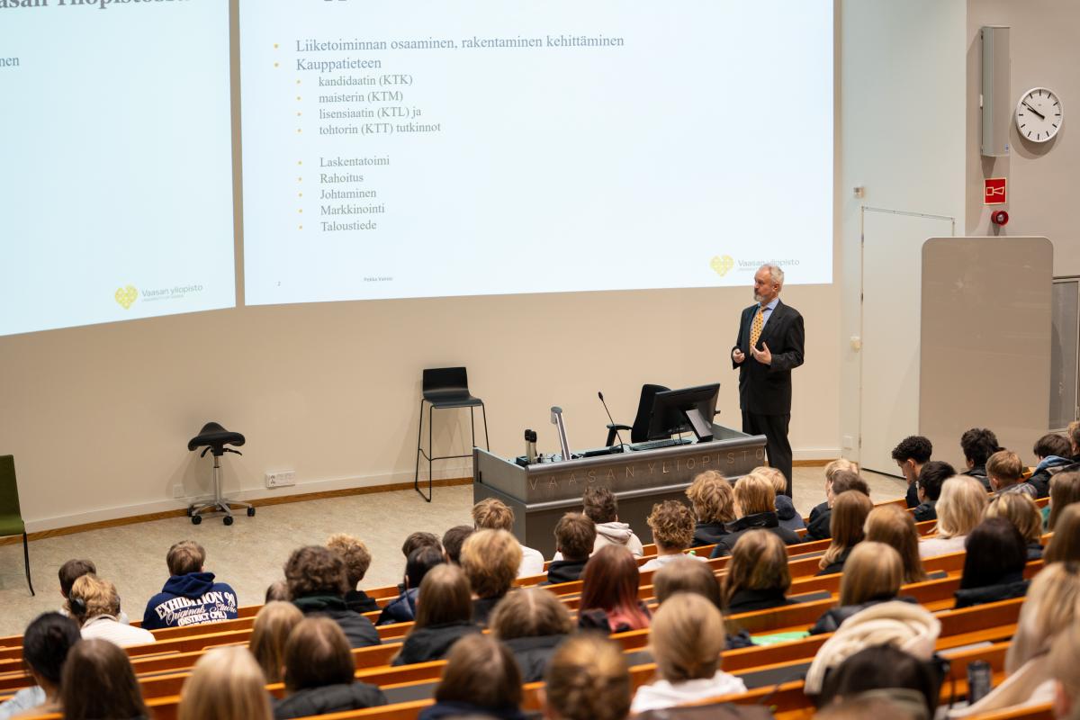 Henkilö luennoimassa auditorion edessä. Katsomo on täynnä ihmisiä ja he kuuntelevat luennoitsijaa.