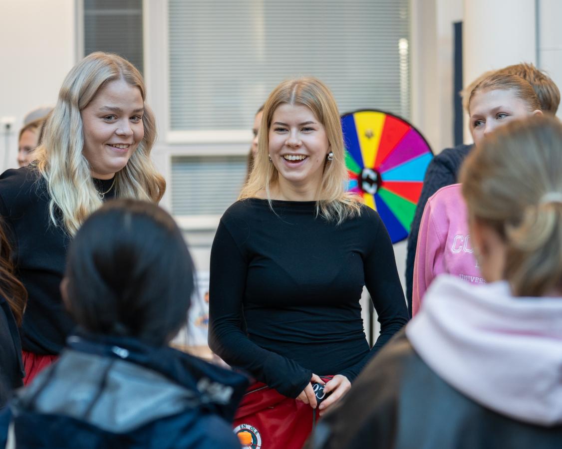 Henkilö punaiset haalarit jalassa ja musta paita päällä. Kasvoillaan hänellä on nauravainen ilme.