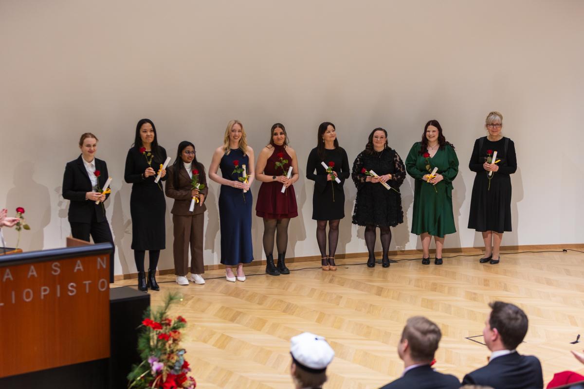 Joukko valmistuneita seisomassa auditorion lavalla käsissään muistamiskääröt ja punaiset ruusut. Kaikki katsovat yleisöön päin ja hymyilevät.