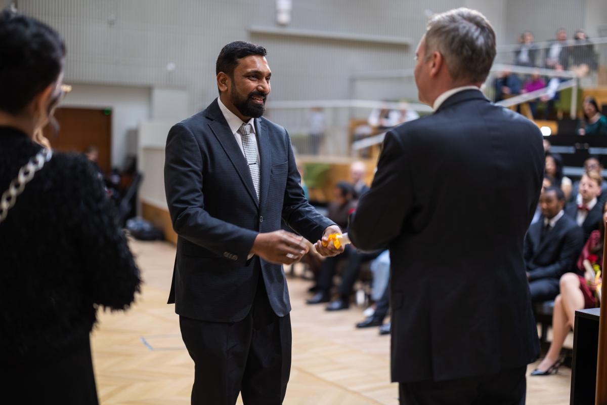 Dekaani ojentamassa muistamiskääröä valmistuneelle. He ovat juuri kättelemässä ja valmistuneella on kasvoillaan iso hymy.