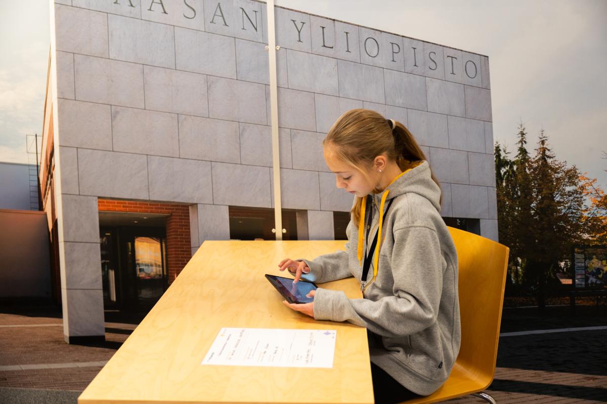 Nuori käyttää tablettia, taustalla kuva Vaasan yliopiston kampuksesta. Kuva: Jonna N. Photography / Nuorten yrittäjyys ja talous NYT