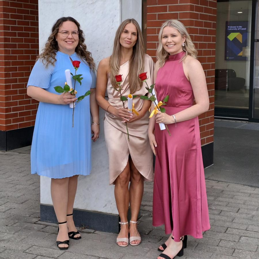 Kolme henkilöä seisomassa juhlavaatteet päällä. Kaikki katsovat kameraan ja hymyilevät leveästi. Jokaisella on käsissään paperinen käärö sekä ruusu.