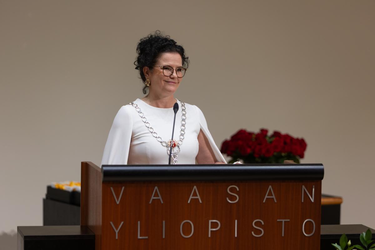 Henkilö Vaasan yliopiston puhujanpöntössä. Päällään hänellä on valkoinen mekko sekä kaulassa hopeinen yliopiston koru.
