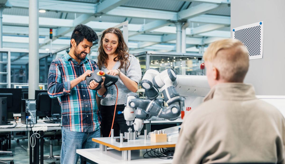 Kolme henkilöä seisomassa tehdastilassa. Kaksi henkilöä miettivät miten robottikäsi saadaan toimimaan. Kolmas henkilö istuu selkä kameraan päin ja seuraa heidän tekemistään.