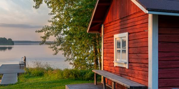 Mökkimaisema, jossa sauna on etualalla ja taustalla näkyy järvi.