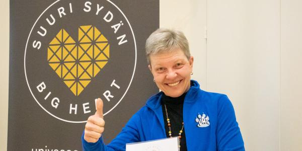 Tiina Jokinen peukuttaa hymyillen Vaasan yliopiston Suuri Sydän -bannerin edessä.