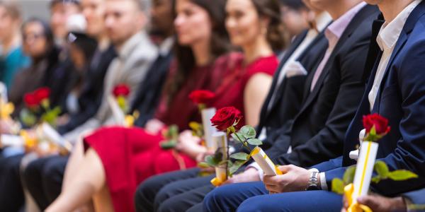 Valmistuneita opiskelijoita istumassa audiotriossa. Kuvan fokus on keskittnyt mieshenkilön kädessä olevaan ruusuun sekä muistaiskääröön. Taustalla näkyy sumeana muita henkilöitä.