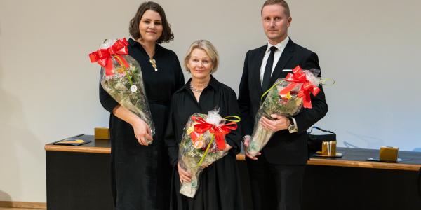 Kolme henkilöä seisomassa kukka-asetelmat käsissään. Kaikki katsovat kameraan ja heillä on päällään juhlavaatteet.