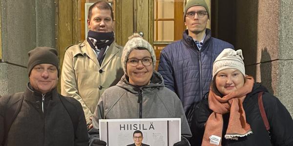 Viisi henkilöä seisomassa rakennuksen portailla kahdessa eri rivissä. Kaikki katsovat kameraan ja hymyilevät. Eturivissä keskellä olevalla henkilöllä on kädessään paperi, jossa on tekoälyllä luotu opettajan kuva kaikista portailla seisovista henkilöistä.