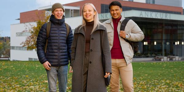 Kolme henkilöä seisomassa Vaasan yliopiston Ankkuri-rakennuksen edessä. Taustalla näkyy rakennus ja pilvinen sinitaivas.