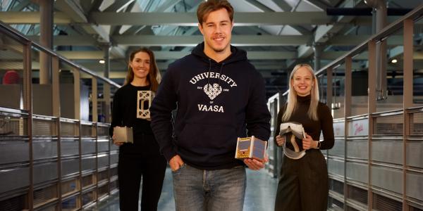 Kolme henkilöä seisomassa käytävällä. Jokaisella on käsissään jokin esine ja kaikki katsovat kameraan hymyillen.