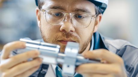 A man with safety glasses and hat on looking closely at two metal components