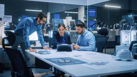 Three people huddled over a table with a laptop and technical drawings in a modern office 