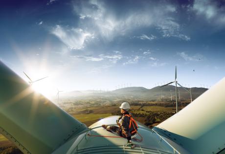A worker sitting on the top of a wind turbine looking at the sun shining on a blue sky