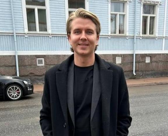 Mieshenkilö seisomassa tien vieressä. Hän poseeraa kameraan päin ja hymyilee. Kuvan taustalla näkyy sininen puutalo sekä mustan auton etuosa.