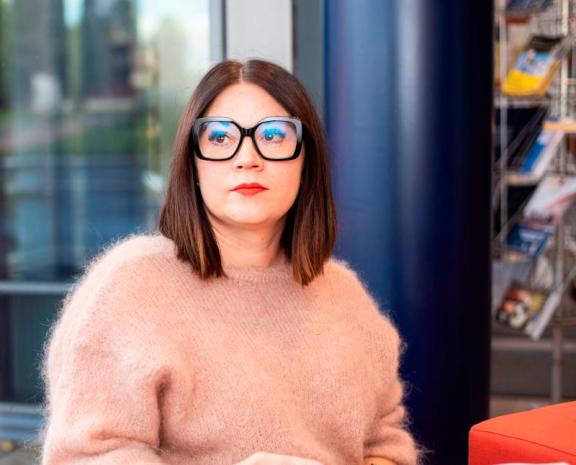 Naishenkilö istumassa pienellä sohvalla. Taustalla näkyy sumeana ikkuna. Naisella on päällä roosan värinen pörröinen neule ja silmillään hänellä on mustasankaiset silmälasit. Käsissään hän pitää kirjaa.