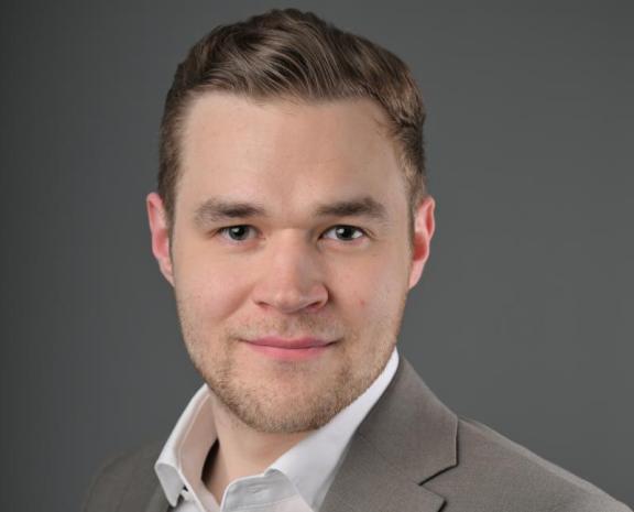 Mieshenkilö katsoo kameraan kasvoista otetussa lähikuvassa. Päällään hänellä on valkoinen kauluspaita ja ruskeanharmaa puvuntakki.