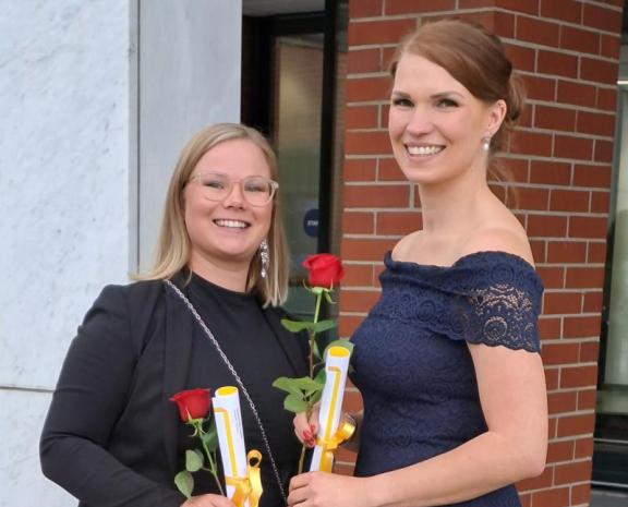 Kaksi henkilöä seisomassa juhlavaatteet päällä pihalla. Molemmat poseeraavat kameralle ja hymyilevät leveästi. Käsissään heillä molemmilla on paperikäärö sekä ruusu.