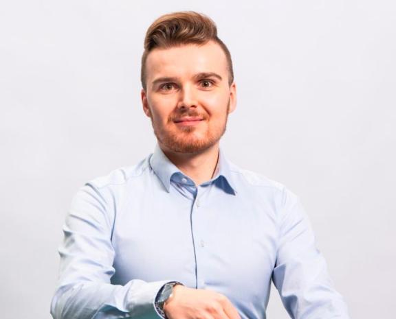 Mieshenkilö istumassa valkoisella tuolilla ja poseeraamassa kameraan. Hänellä on päällään vaalean sininen kauluspaita ja tummansiniset siistit housut. Hiukset ja silmät ovat ruskean väriset. Hän katsoo kohti kameraa ja hymyilee.