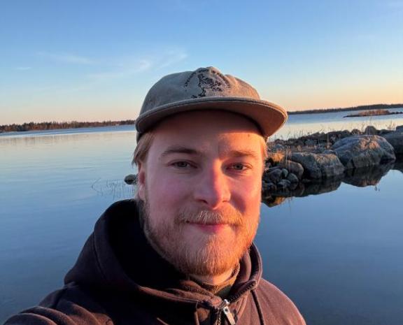 Mieshenkilö seisomassa meren rannassa. Hän poseeraa kameraa kohti ja taustalla näkyy tyyni vesi sekä kirkas sininen taivas-